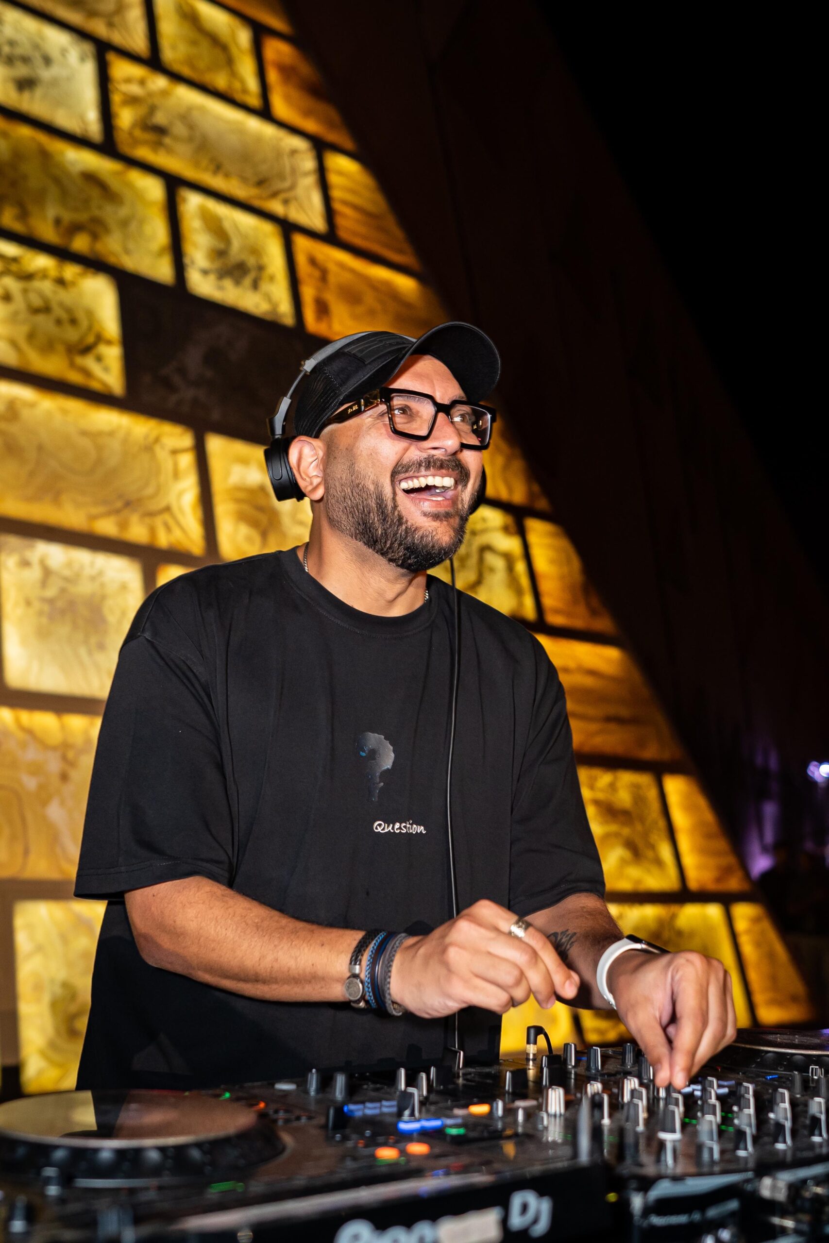 Saify smiling behind DJ decks, wearing headphones, glasses and a black cap.