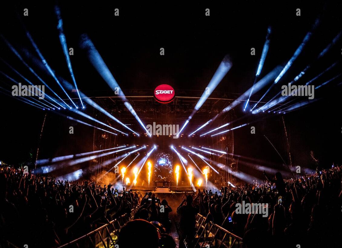 Sziget festival crowd facing a stage with white spotlights and flame effects.